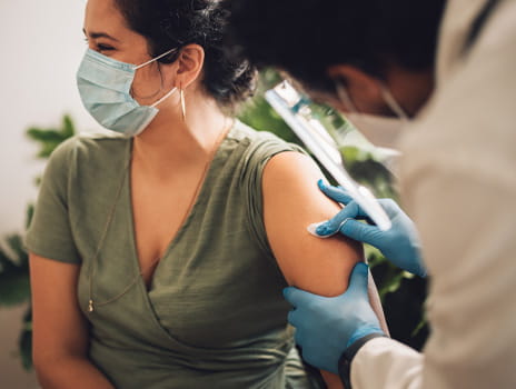 Woman wearing a face mask looking away as a healthcare provider cleans her upper arm to prepare for a vaccination. 