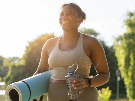 Smiling woman carrying a yoga mat and water bottle on a sunny day