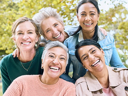 Five smiling women of various ages and ethnicities are grouped together looking at the camera in front of a tree-filled sky.